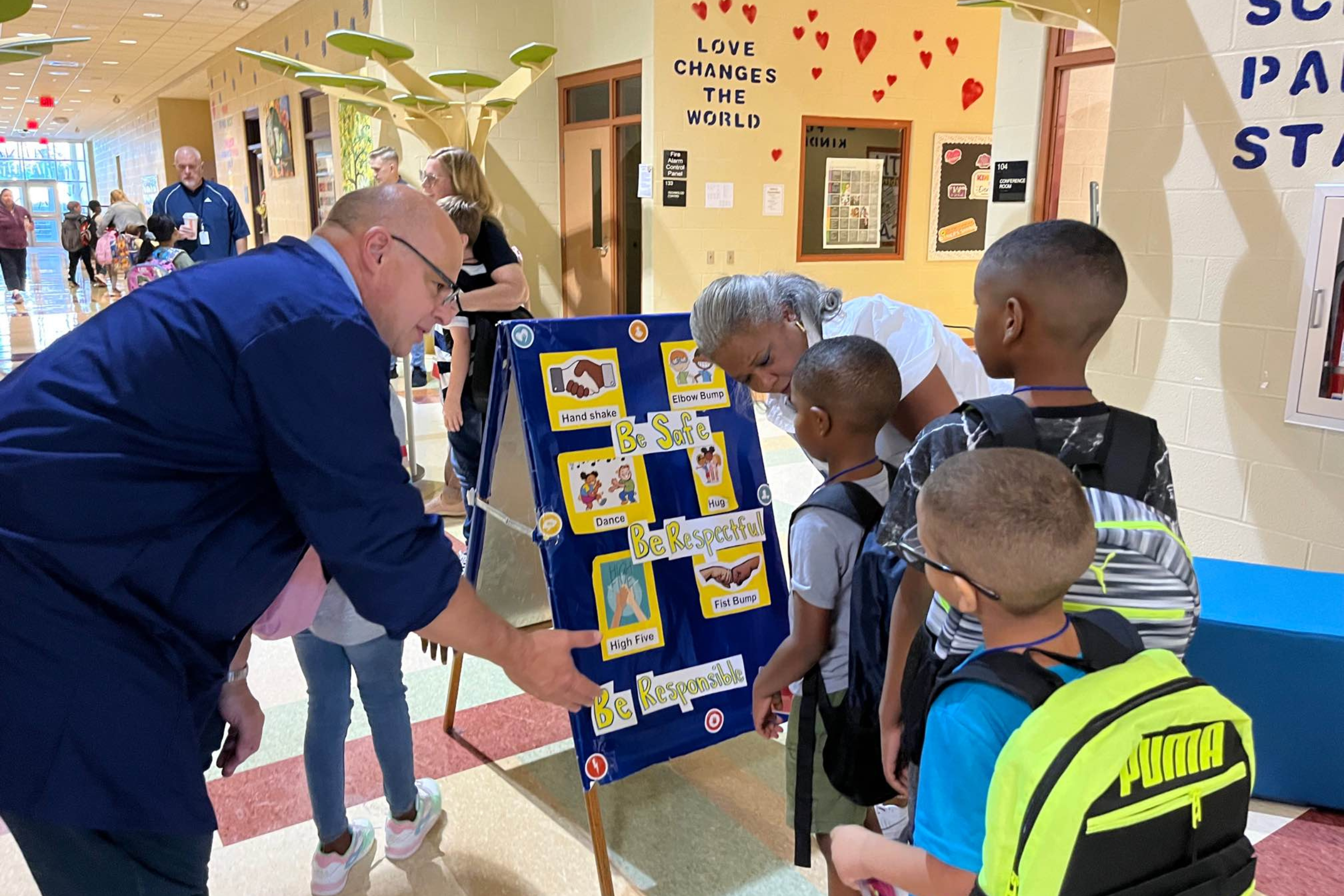 Principal and teacher greet students before walking into their classroom with an option board for choosing what kind of greeting they would like. They can choose from high fives, waves, or fist bumps.