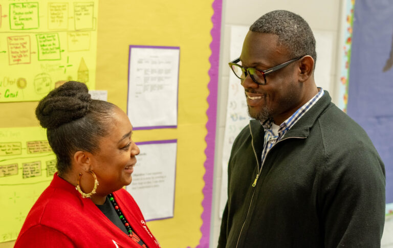 Black male principal standing in a school hallway smiling at a female educator.